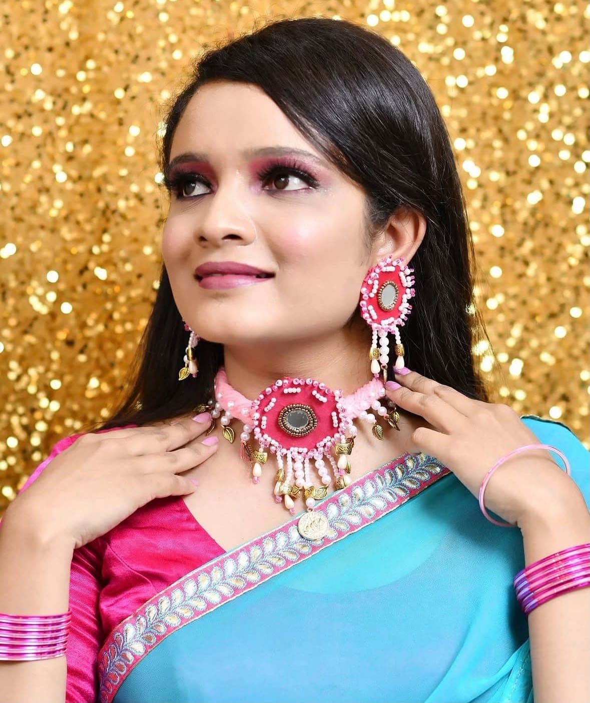 A woman in a pink beaded choker and matching earrings, hands raised to her collar, in front of a gold sparkle backdrop