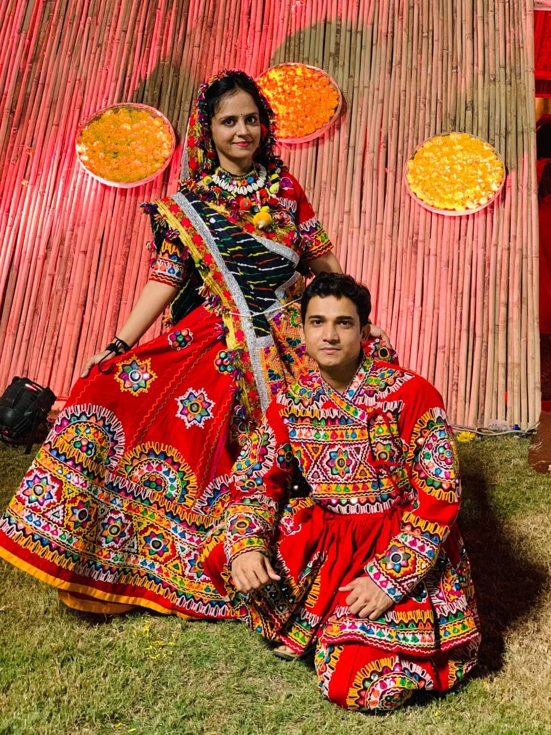 A man and a woman in red embroidered Navratri outfits, posed on a lawn in front of a bamboo wall