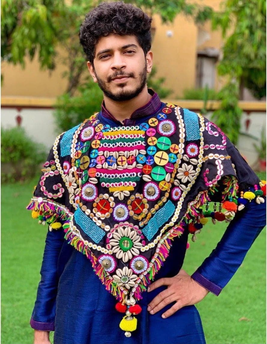 A man in a black poncho covered with buttons, cowrie shells, and tassels, over a blue kurta