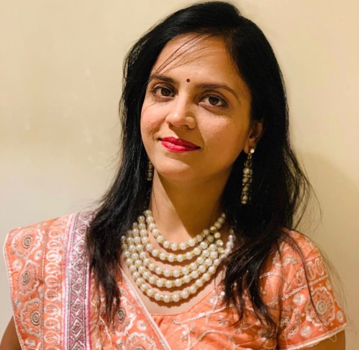 A woman with long black hair in an orange embroidered outfit and a layered pearl necklace, in front of a cream wall