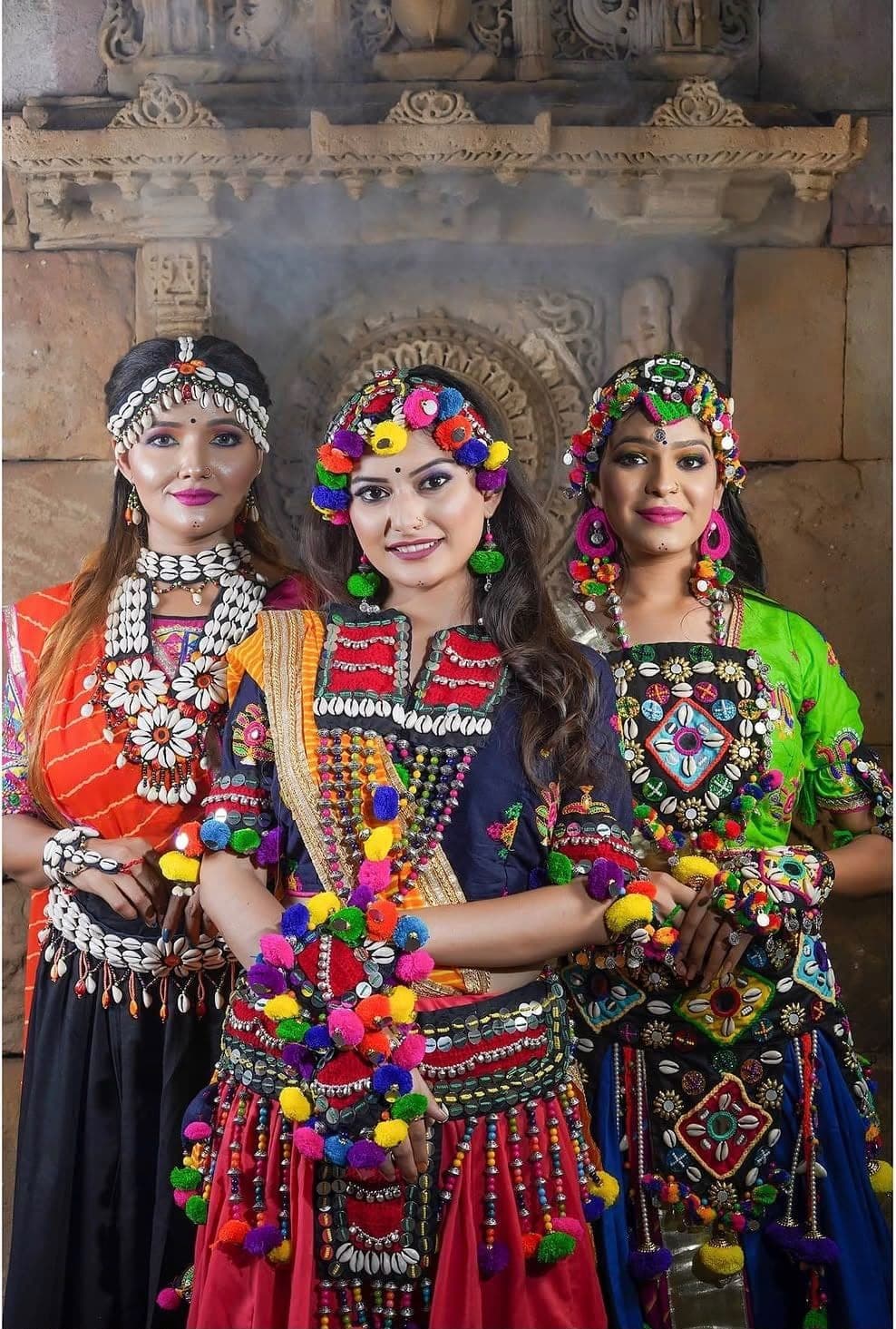 Three women in colourful Navratri jewellery, standing in front of a carved stone wall