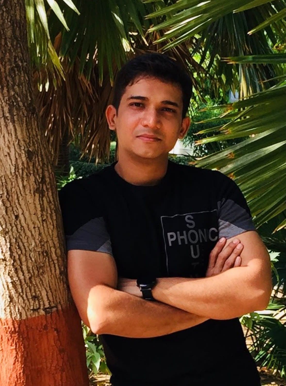 A man in a black t-shirt leaning against a tree trunk with his arms crossed, palm leaves behind him