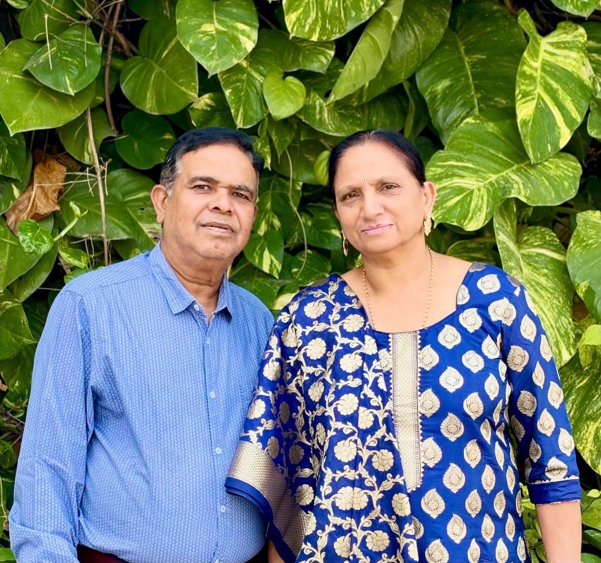 A man in a blue shirt and a woman in a blue and gold suit, standing together in front of a wall of green leaves